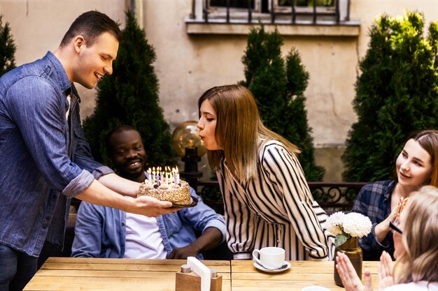 Tarta de cumpleaños con velas encendidas que la niña está apagando y mejores amigas en la terraza de un acogedor café