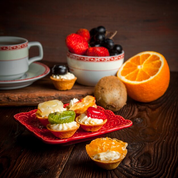 Tartaletas variadas con taza de té y bandeja de frutas y naranja en plato