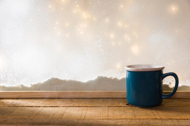 Taza azul en la mesa de madera cerca del banco de nieve y luces de hadas