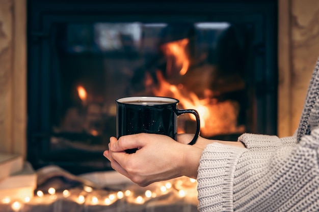 Foto gratuita una taza con una bebida caliente en manos femeninas sobre un fondo borroso de chimenea