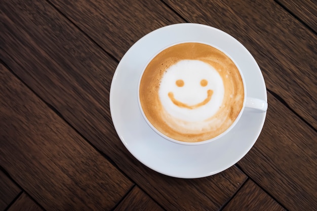 Taza blanca de la cara feliz de la sonrisa del arte del latte en fondo de madera marrón de la tabla.