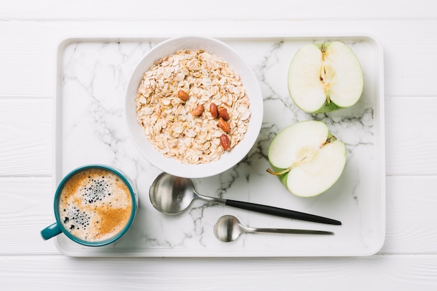 Taza de cafe; avena y manzana a la mitad con cucharas en bandeja sobre mesa