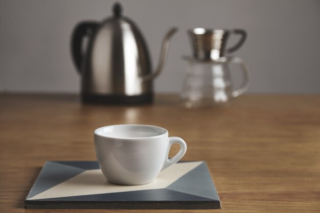 Taza de café en blanco blanco frente a la tetera moderna y hermosa cafetera de goteo transparente. Taza en placa de cerámica sobre mesa de madera gruesa en cafetería.