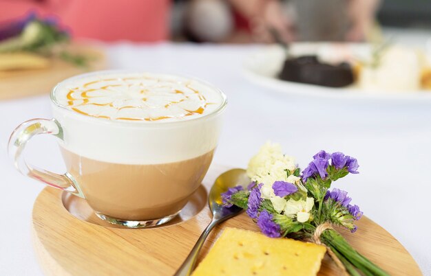Taza de café caliente bien decorada que se sirve en un plato de madera y una pequeña flor violeta y galletas