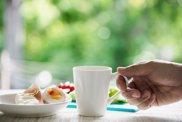 Taza de café caliente con capuchón de la mano con huevos cocidos y ensalada de cebolla y papa con pepino y desayuno con bosque verde - concepto de comida de desayuno