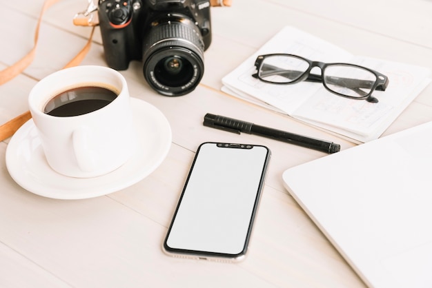 Taza de café; cámara; Teléfono móvil; bolígrafo; gafas en el cuaderno sobre la mesa de madera