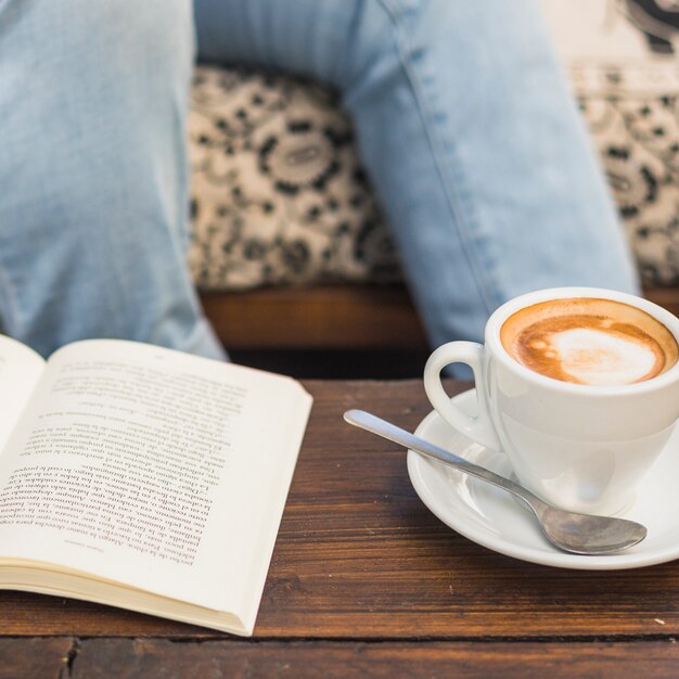 Taza de café capuchino caliente y libro abierto en la mesa de madera con una persona sentada detrás