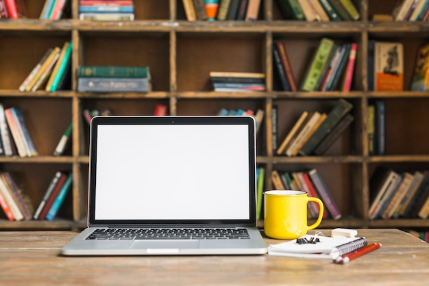 Taza de café y computadora portátil con papelería en el escritorio de madera en la biblioteca