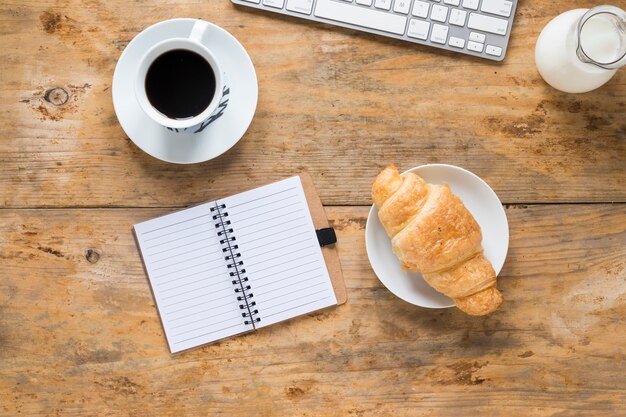 Taza de café; croissant al horno Leche con teclado y bloc de notas en espiral en el escritorio de madera