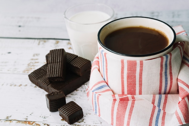 Foto gratuita taza de café envuelta con la servilleta; barra de chocolate oscuro y leche en la mesa de madera