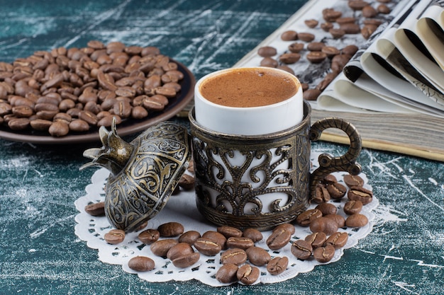 Taza de café espumoso, plato de granos de café y libro sobre mesa de mármol.