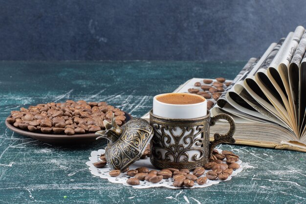 Taza de café espumoso, plato de granos de café y libro sobre mesa de mármol.