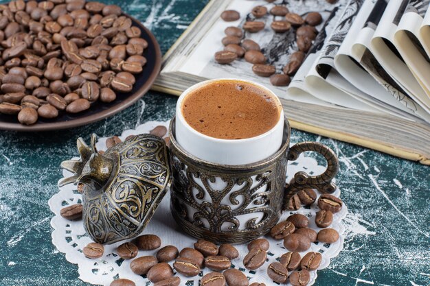 Taza de café espumoso, plato de granos de café y libro sobre mesa de mármol.