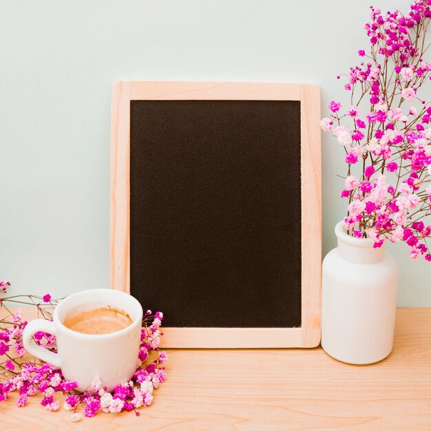 Taza de café y florero rosa para el aliento del bebé cerca de la pizarra de madera en blanco en el escritorio contra la pared