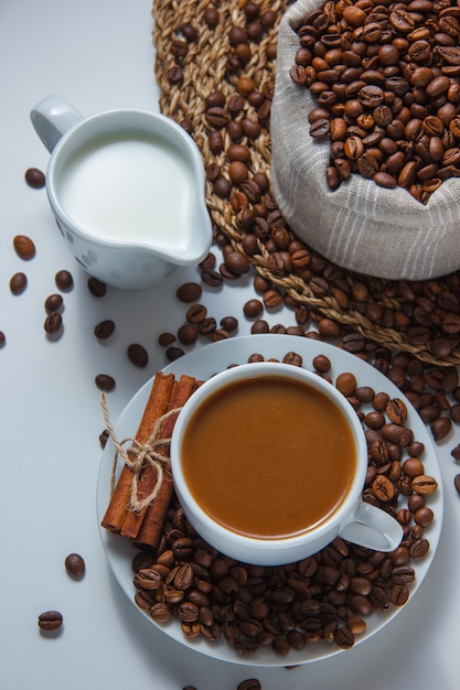 Una taza de café con granos de café en un saco y platillo, leche, canela seca, vista de ángulo alto sobre un salvamanteles y superficie blanca