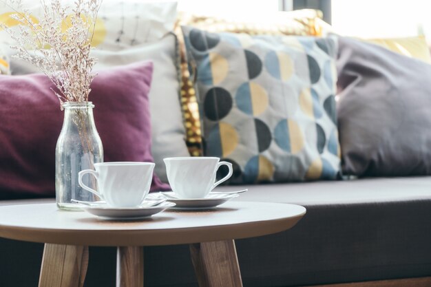 Taza del café con leche con el florero en la decoración de la tabla con la almohada en el sofá