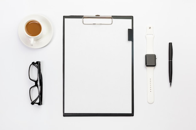 Taza de café, lentes, reloj inteligente, bolígrafo y portapapeles de papel en blanco en el escritorio blanco