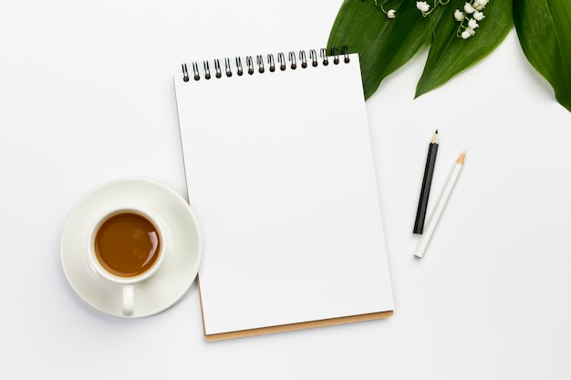 Taza de café, libreta de espiral en blanco y lápices de colores con hojas y flores en el escritorio de oficina