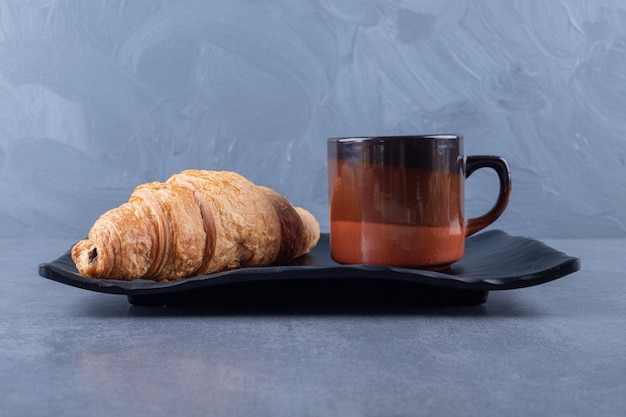 Taza de café negro y croissant para desayunar sobre fondo gris.