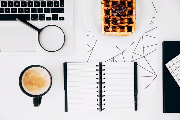 Taza de café; ordenador portátil; lupas; Bloc de notas en espiral y waffles con chocolate en un plato contra el escritorio blanco