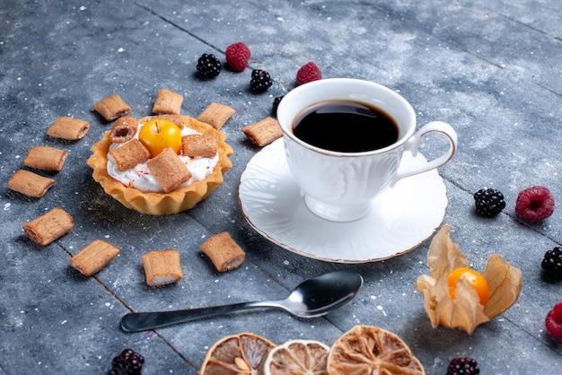 Taza de café con pastel cremoso, forma de almohada, galletas junto con bayas en el escritorio gris, galleta de baya