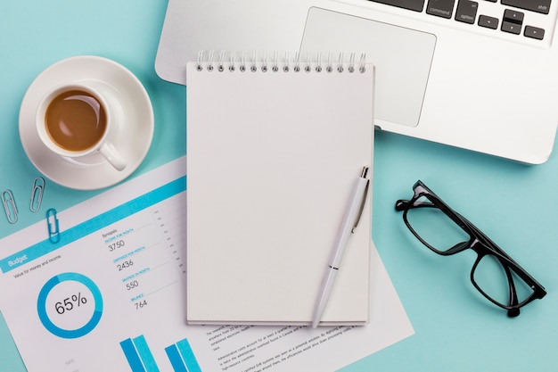 Taza de café, plan de presupuesto, bloc de notas en espiral, bolígrafo, lentes y computadora portátil sobre fondo azul