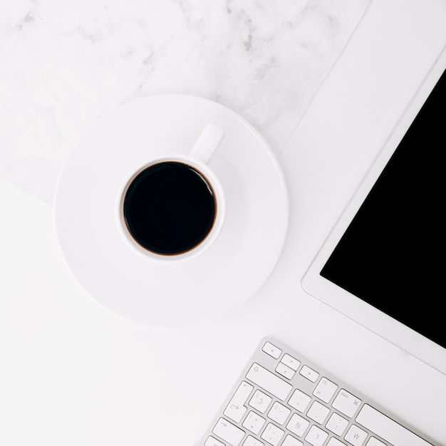 Taza de café en el platillo cerca de la tableta digital y el teclado en el escritorio blanco