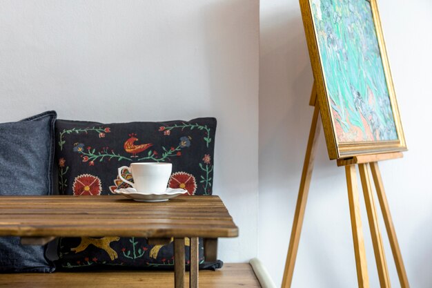 Taza de café y platillo en mesa de madera frente a cojín y caballete