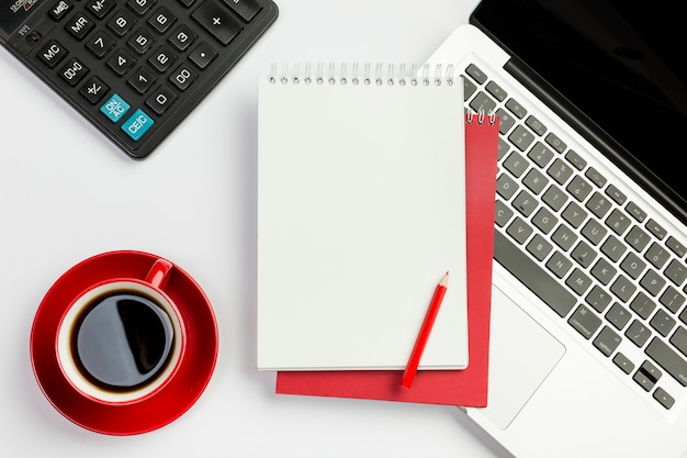 Taza de café rojo, calculadora, bloc de notas en espiral, lápiz en la computadora portátil sobre el fondo blanco