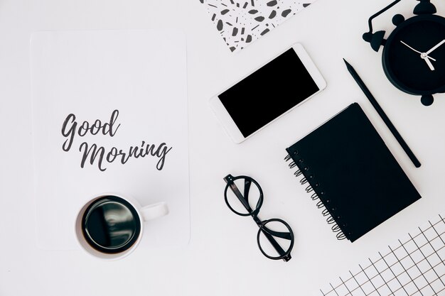 Taza de café sobre papel con texto de buena mañana; Teléfono móvil; Despertador y papelería en escritorio blanco