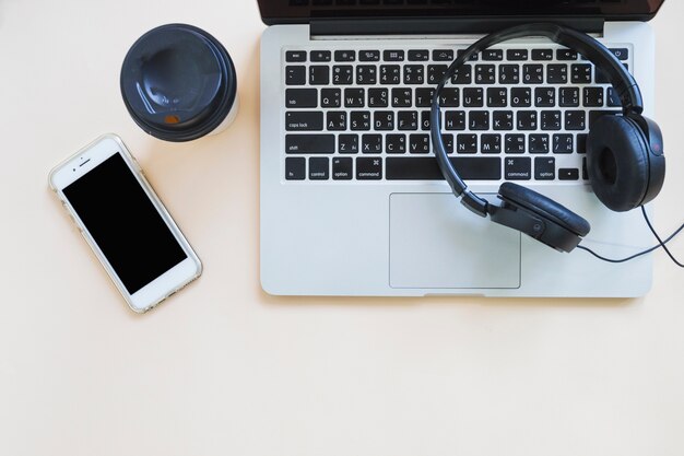 Taza de café; teléfono móvil y auriculares en la computadora portátil contra el fondo beige