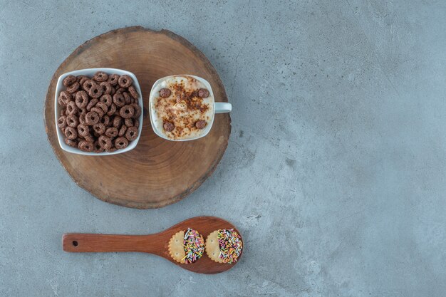 Una taza de capuchino y un cuenco de maíz aro en un tablero junto a la cuchara, sobre el fondo azul.