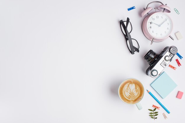 Taza de capuchino con latte art; camara vintage despertador; Lápiz y lentes sobre fondo blanco