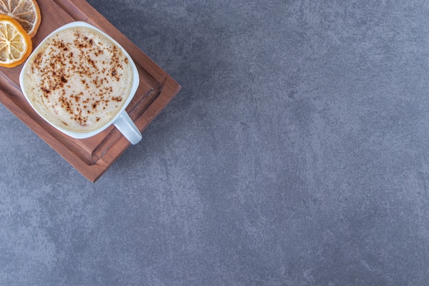 Una taza de capuchino en la placa de madera junto a rodajas de limón, sobre la mesa azul.