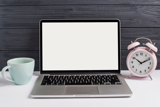 Taza de cerámica; Despertador y computadora portátil abierta en el escritorio blanco sobre fondo negro de madera