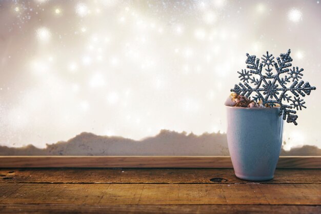 Taza con copo de nieve de juguete en la mesa de madera cerca del banco de nieve y luces de hadas