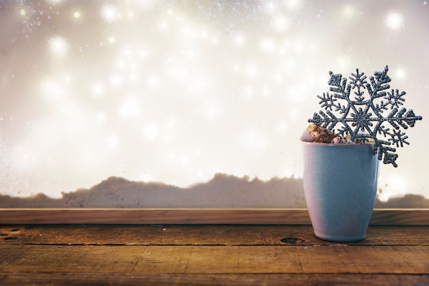 Taza con copo de nieve de juguete en la mesa de madera cerca del banco de nieve y luces de hadas