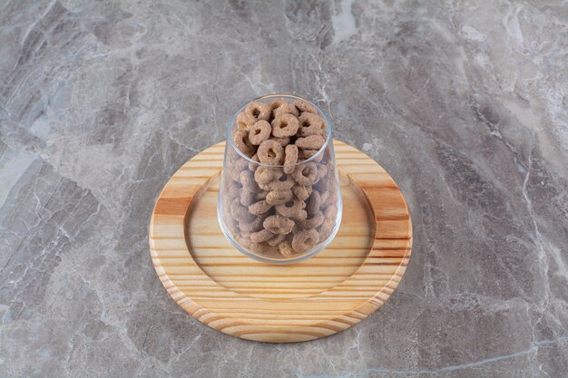 Una taza de cristal llena de anillos de cereal de chocolate sobre una tabla de madera.