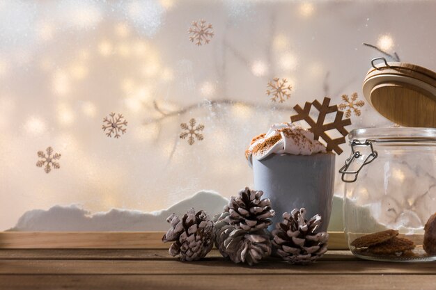 Taza con ganchos y lata en una mesa de madera cerca del banco de nieve, planta ramita, copos de nieve y luces