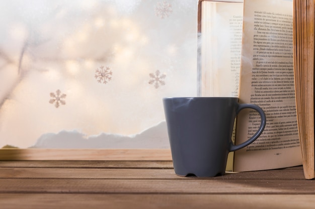 Taza y libro en mesa de madera cerca de banco de nieve y copos de nieve