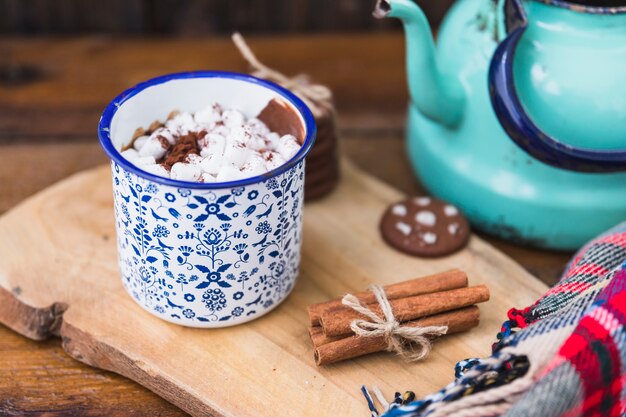 Taza con malvavisco cerca de galletas, canela y hervidor