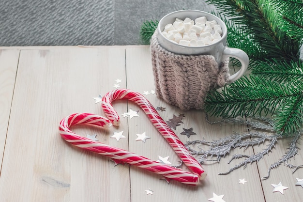 Taza de malvaviscos y bastones de caramelo rodeados de adornos navideños en una mesa de madera