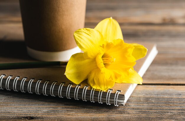 Taza de papel desechable portátil y flor amarilla sobre un fondo borroso de madera