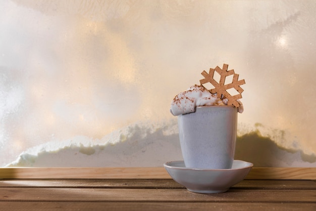 Taza en un plato con un copo de nieve de juguete en una mesa de madera cerca del banco de nieve