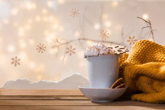 Taza en el plato con galletas cerca de la bufanda en la mesa de madera cerca del banco de nieve y luces de colores