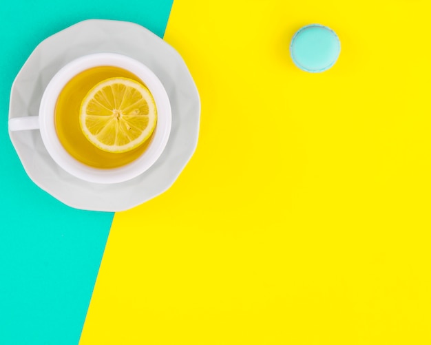 Taza y plato de té de limón blanco con macarrones sobre fondo turquesa y amarillo