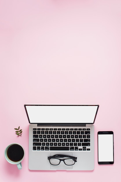 Foto gratuita taza de te los anteojos; teléfono móvil y una computadora portátil abierta con pantalla blanca sobre fondo rosa.