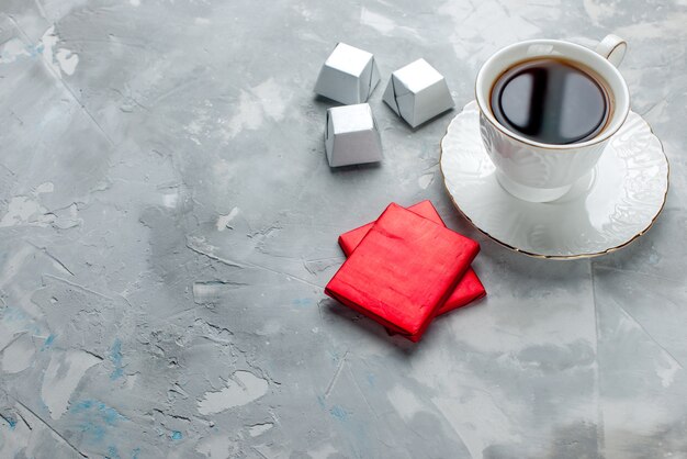 Taza de té caliente dentro de la taza blanca sobre placa de vidrio con paquete de plata, caramelos de chocolate en la mesa de luz, té dulce galleta de chocolate
