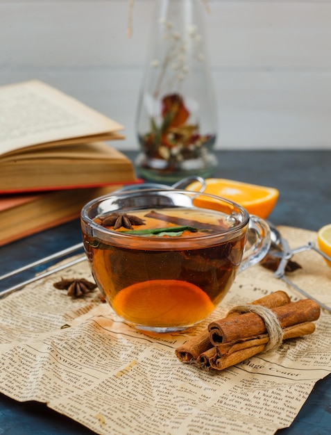 Una taza de té y canela con periódico, naranja y un florero.