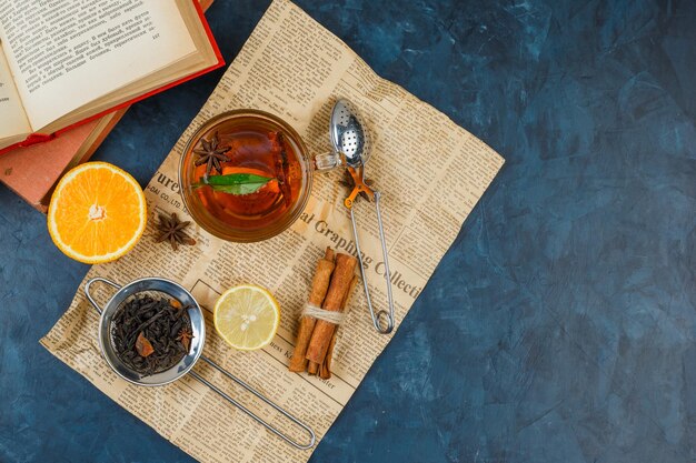 Una taza de té, coladores de té, canela y naranja con periódico y un libro.